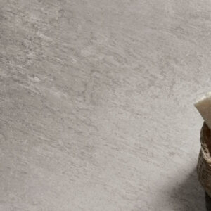 Alpine Sepia light grey quartz-effect porcelain tile swatch displayed alongside a small wicker basket containing sponges, highlighting the neutral stone-coloured texture.