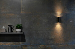 Patina Blue metallic-finish porcelain tiles on a kitchen wall, featuring a weathered deep-teal stone-look surface with copper-coloured oxidised patches, illuminated by a warm downlighter positioned above a minimalist floating shelf.