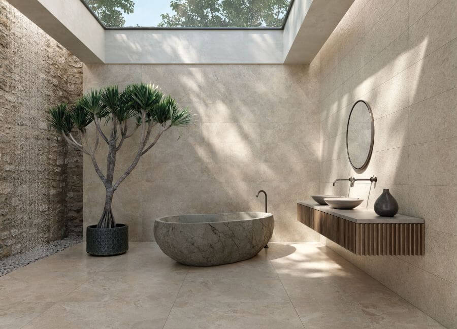 NATURE-STONE-EFFECT-TILES-FOR-BATHROOMS-SAND. Image shows stone effect tiles in bathroom with a stone bath and a large tree and a vanity unit